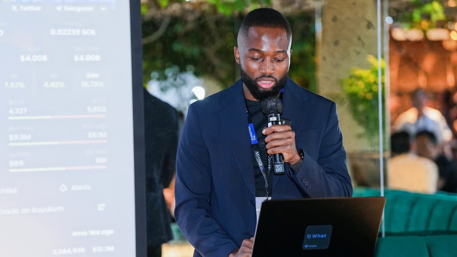 Grey Jabesi looking down at a laptop while speaking at a crypto event, used for an article about AI and trading.